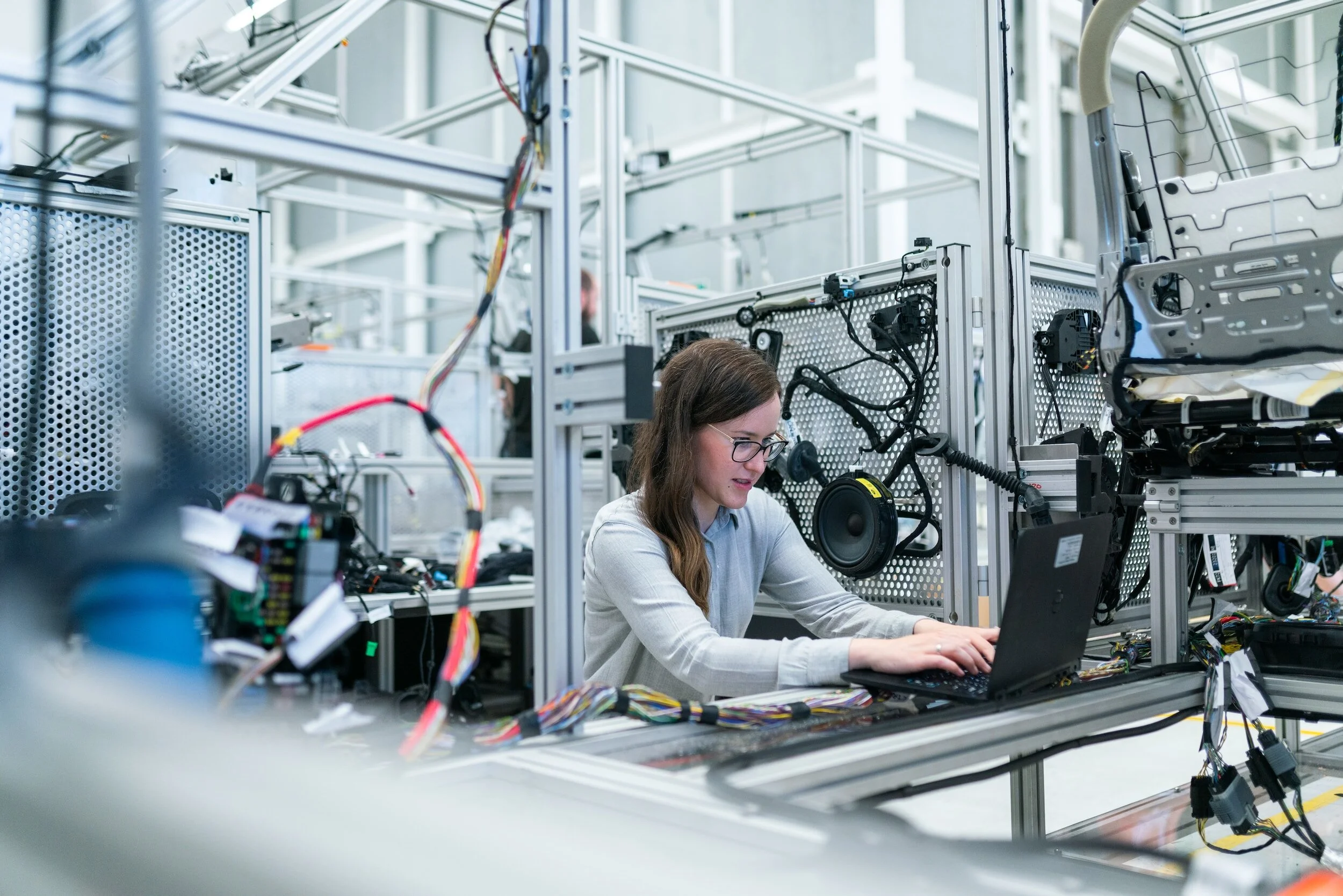 Woman working on a laptop in an industrial setting with electronic equipment, wires, and machinery.