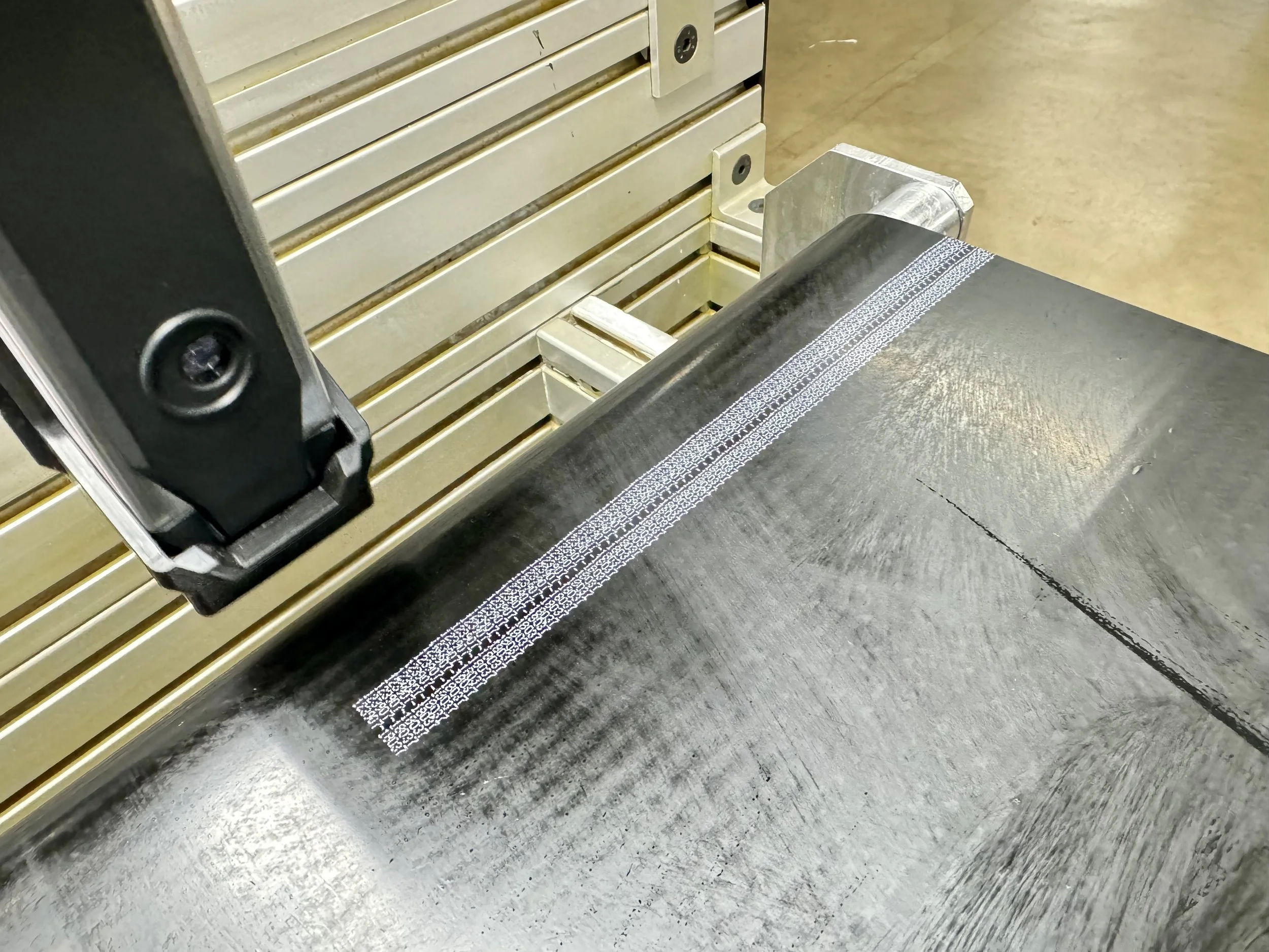 Close-up of a high-precision 3D printer bed with a metallic print, showing detailed printing lines and a partially printed intricate white pattern on the surface.